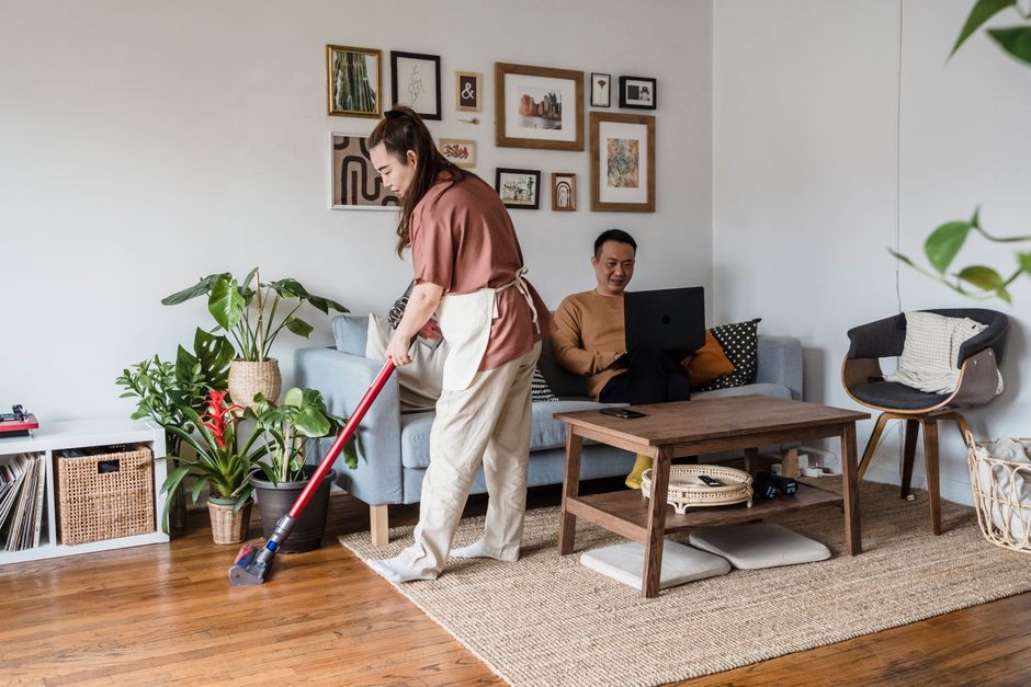 Woman vacuuming living room while man on laptop, modern home setting.