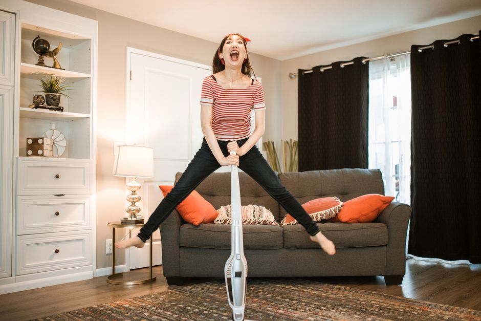 A joyful woman making cleaning fun by jumping with a vacuum in her living room.